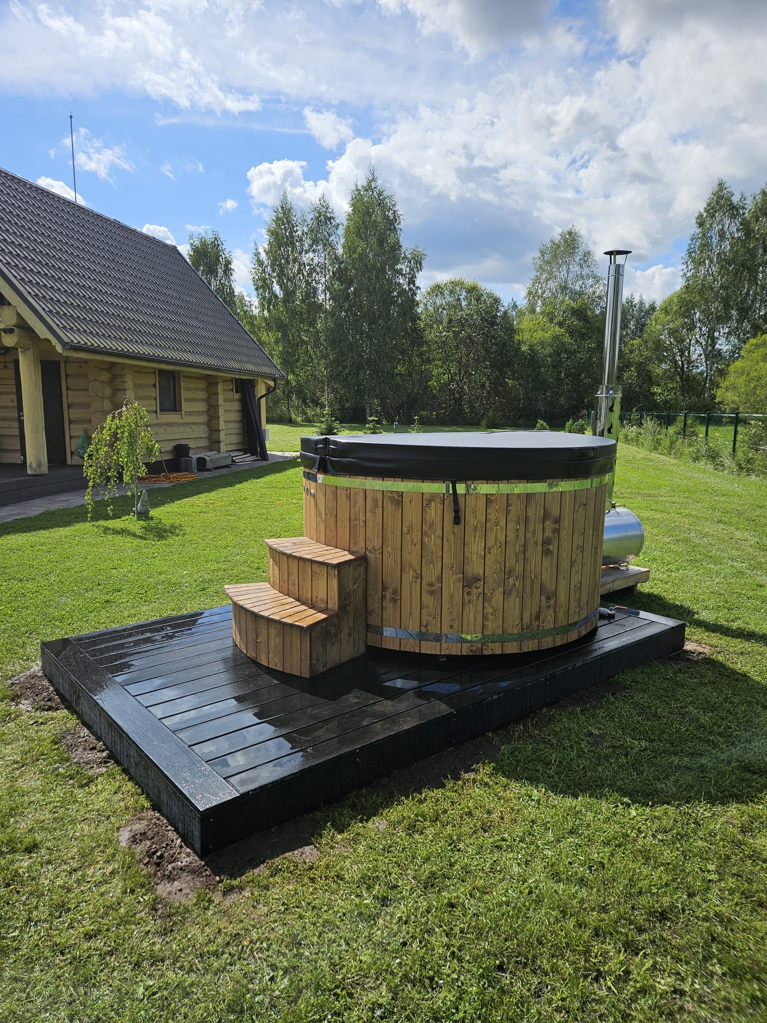 Wooden hot tub on a grassy area with a wooden cabin and trees in the background.