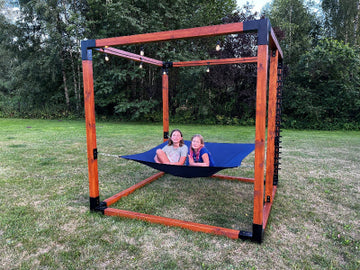 Two children playing in a blue hammock attached to a wooden frame structure in a grassy outdoor area.