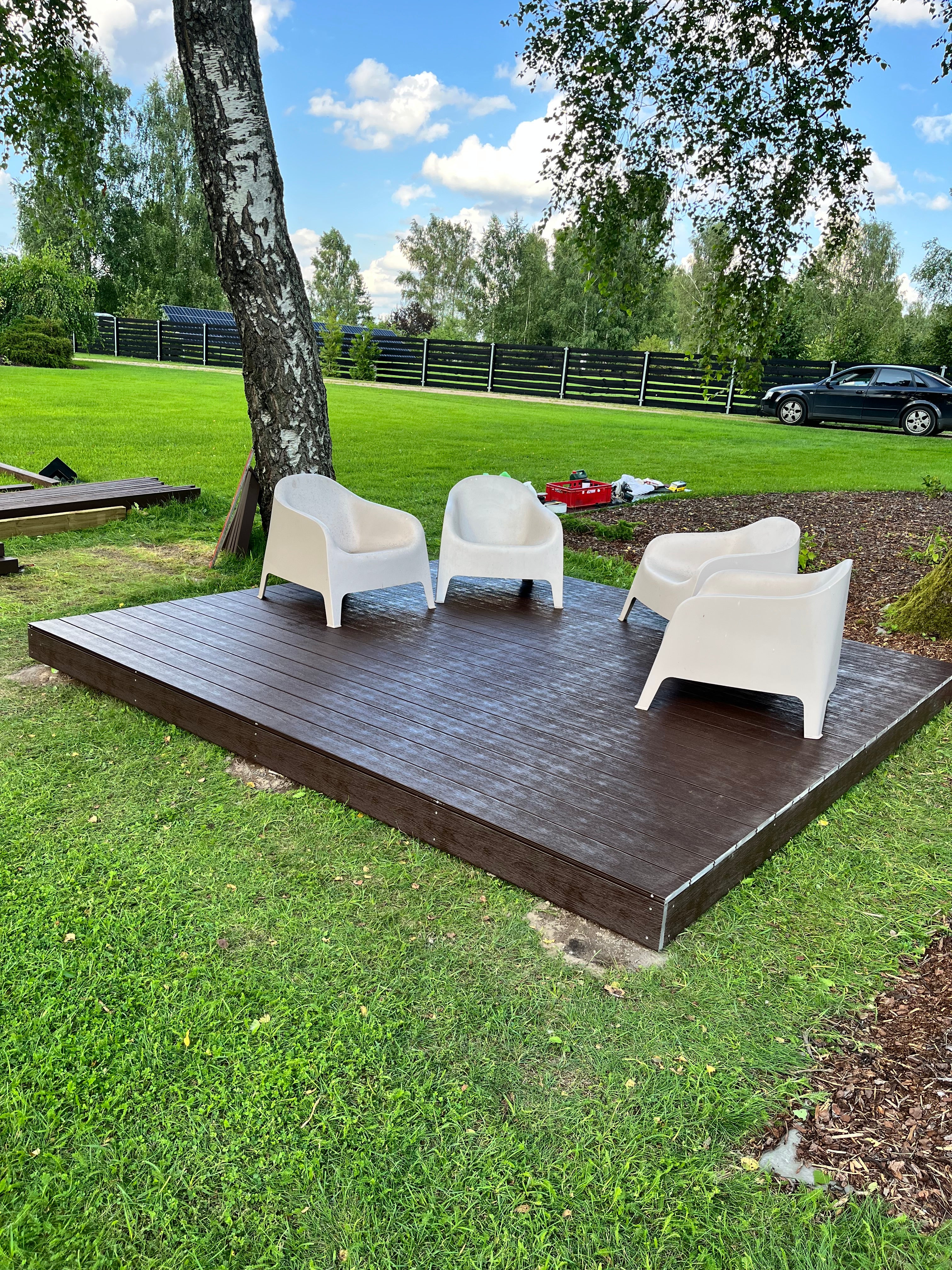 White chairs on a wooden platform in a grassy area with trees and a blue sky.