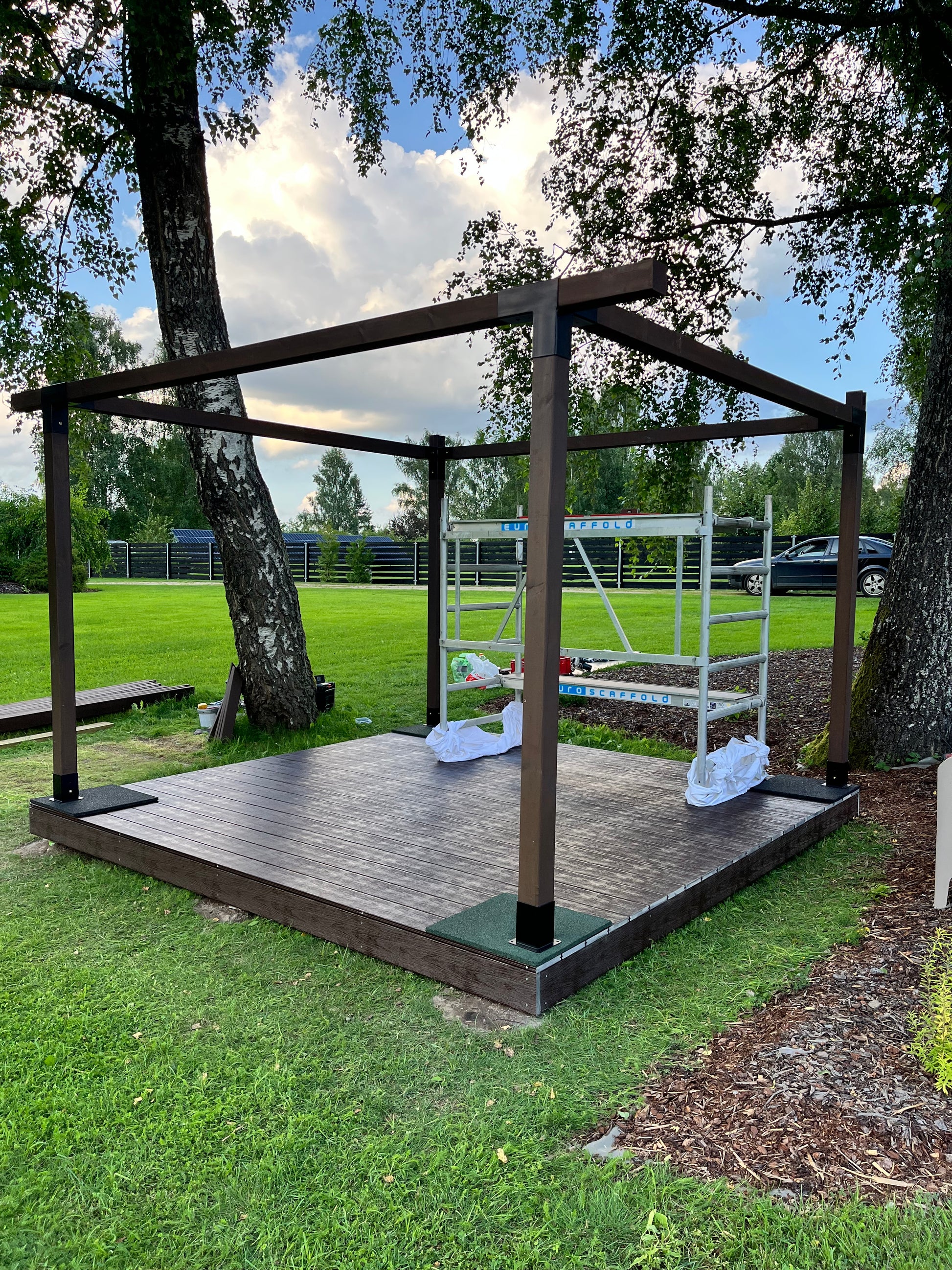 Wooden play structure in a grassy area with trees and a pool in the background