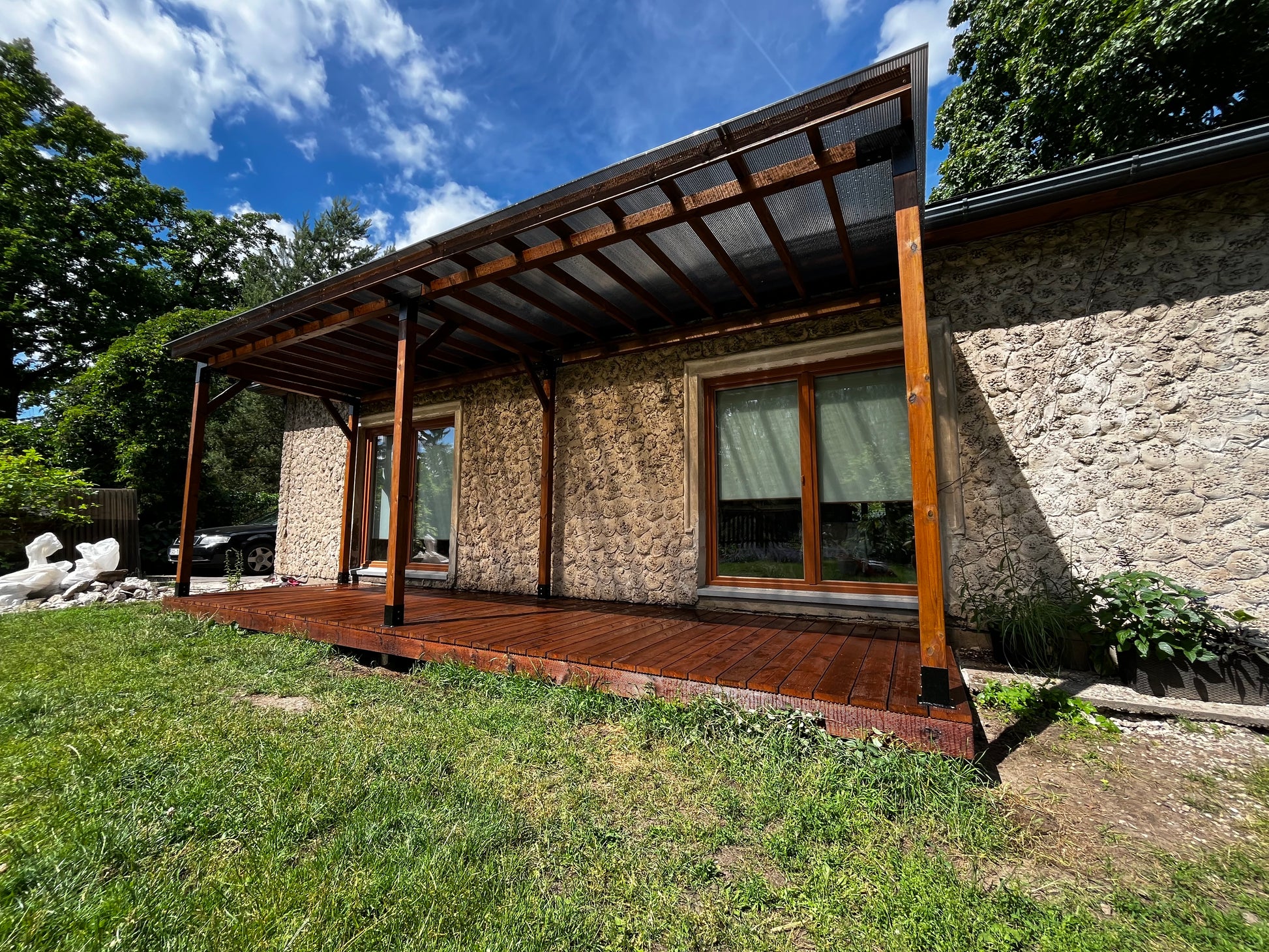 Stone house with a wooden deck and pergola under a blue sky.