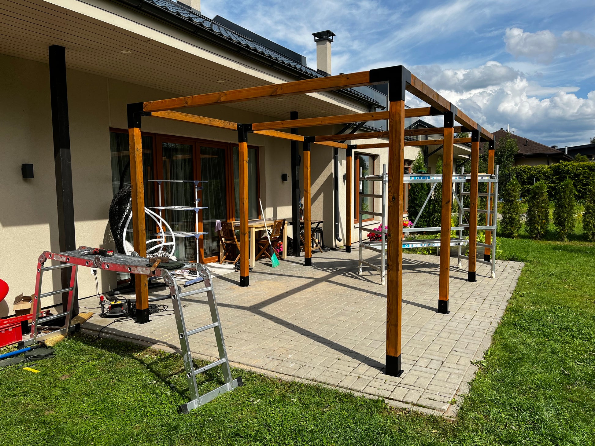 Patio with wooden pergola structure under construction, surrounded by grass and a house.