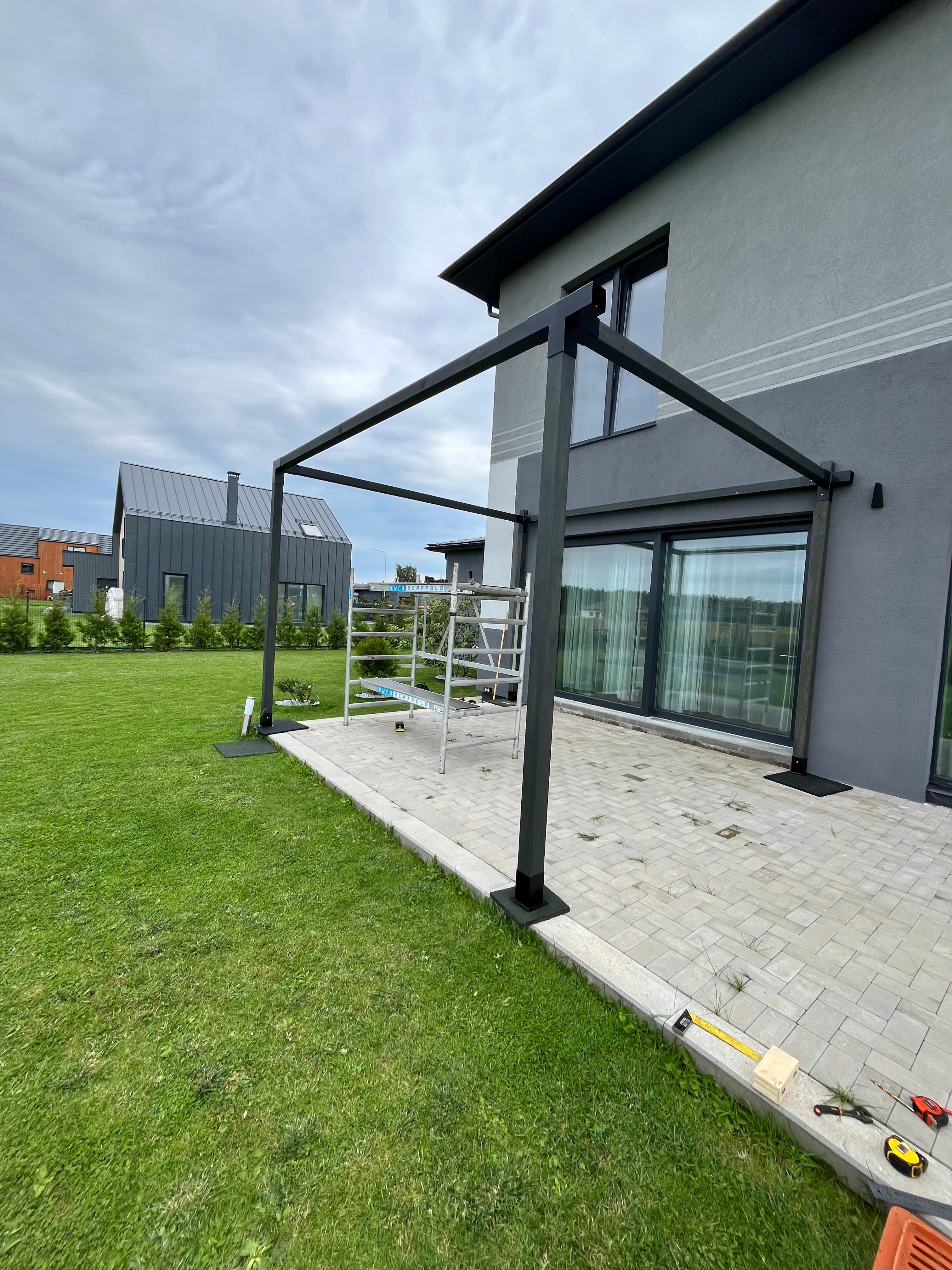 Black metal pergola structure on a grassy area with a modern house in the background