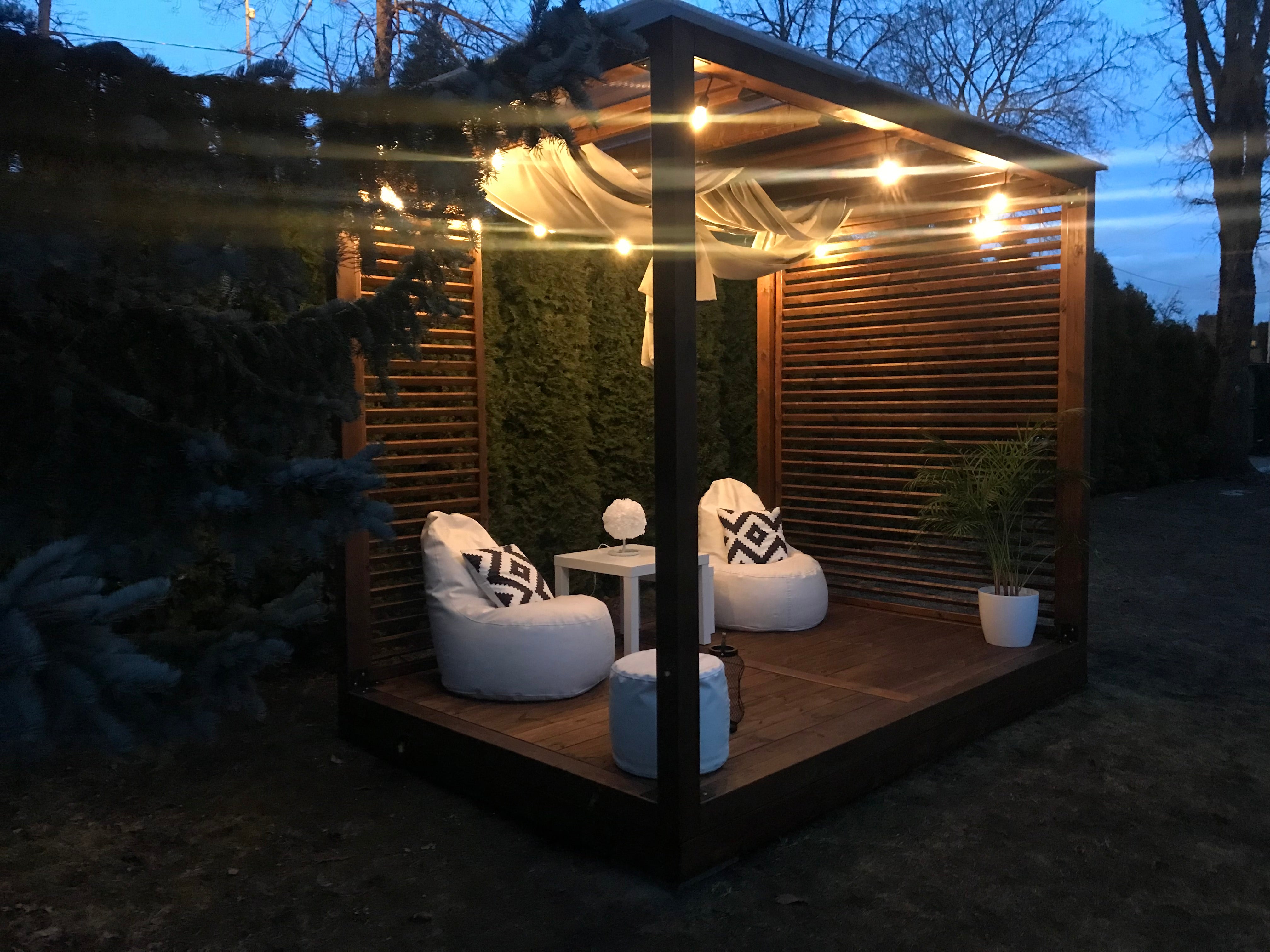Outdoor wooden pavilion with string lights, chairs, and plants at night.