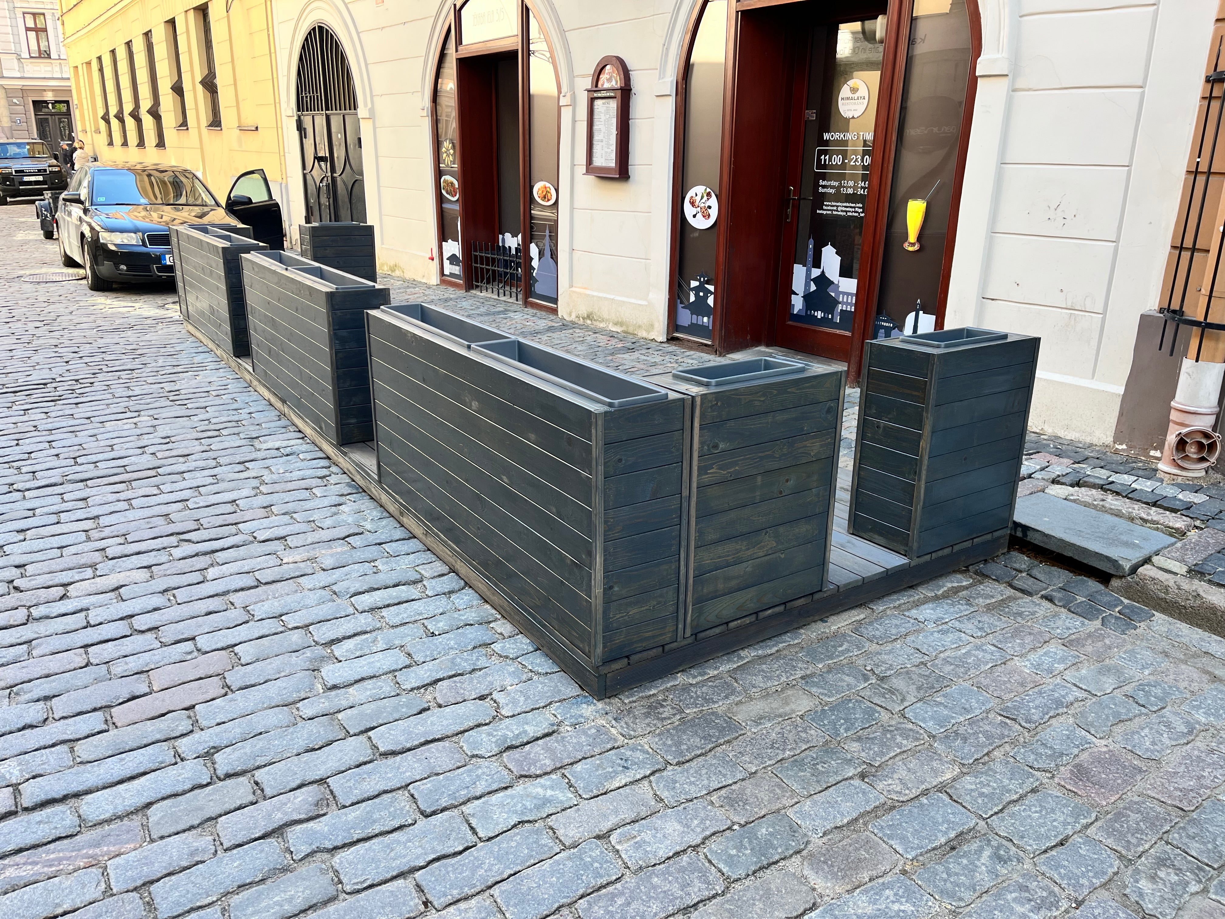Row of black metal planters on a cobblestone street with a building in the background.