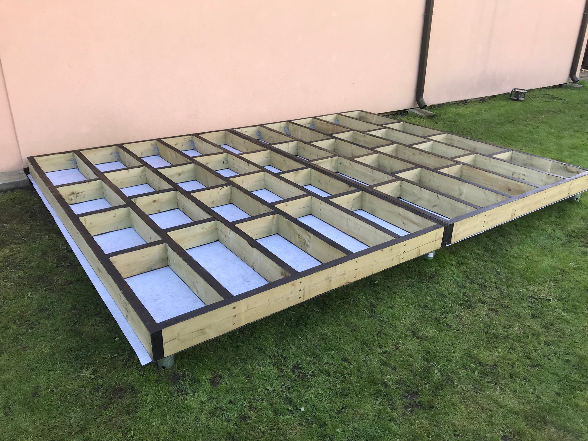 Wooden floor joist system on grass with a beige wall in the background