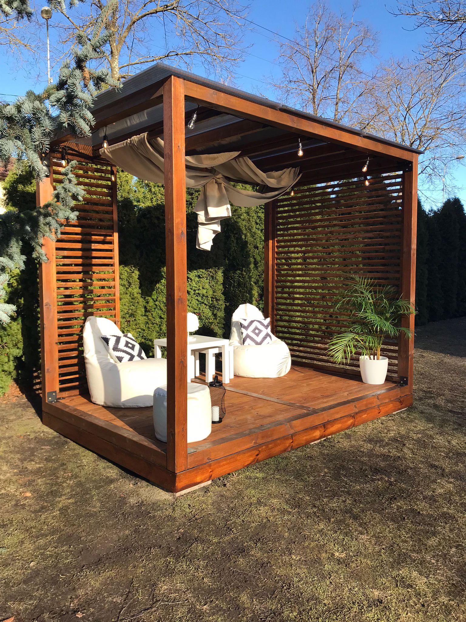 Wooden outdoor gazebo with seating area and plants on a grassy area.